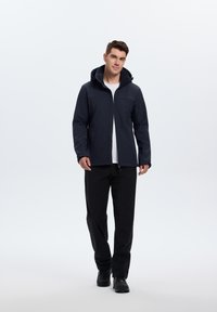 Marineblauwe softshell jas met een ritssluiting aan de voorkant, verstelbare capuchon en contrasterende grijze accenten. Draag het over een wit T-shirt en zwarte broek.