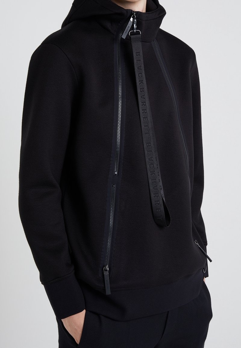 Svart hoodie i mjukt material, med dubbla dragkedjor och präglad logotyp på ett band över framsidan. Texturerade ärmmuddar.