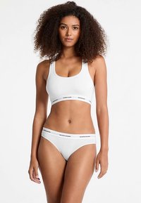 Bralette athlétique blanc et short assorti, présentant une bande épaisse avec le logo "GUESS JEANS". Tissu lisse et extensible, design minimaliste, ajusté à la forme.