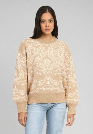 Femme portant un pull ras du cou beige avec un motif floral blanc et un jean bleu clair, debout devant un fond gris uni.