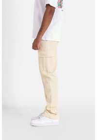 Beige cargo-broeken met meerdere zakken en een label met een patch, gecombineerd met witte sneakers. De stof lijkt zacht en heeft een relaxed fit.