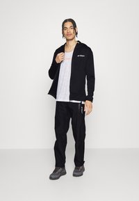 Felpa nera con zip e logo "TERREX", canotta grigia in mesh, pantaloni cargo neri e scarpe da escursionismo grigie con accenti arancioni.