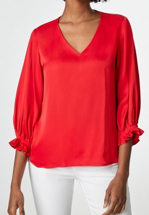 Kobieta w jasnoczerwonej bluzce z dekoltem w kształcie litery V, z marszczonymi rękawami do połowy przedramienia oraz białych spodniach, widziana od szyi do bioder.