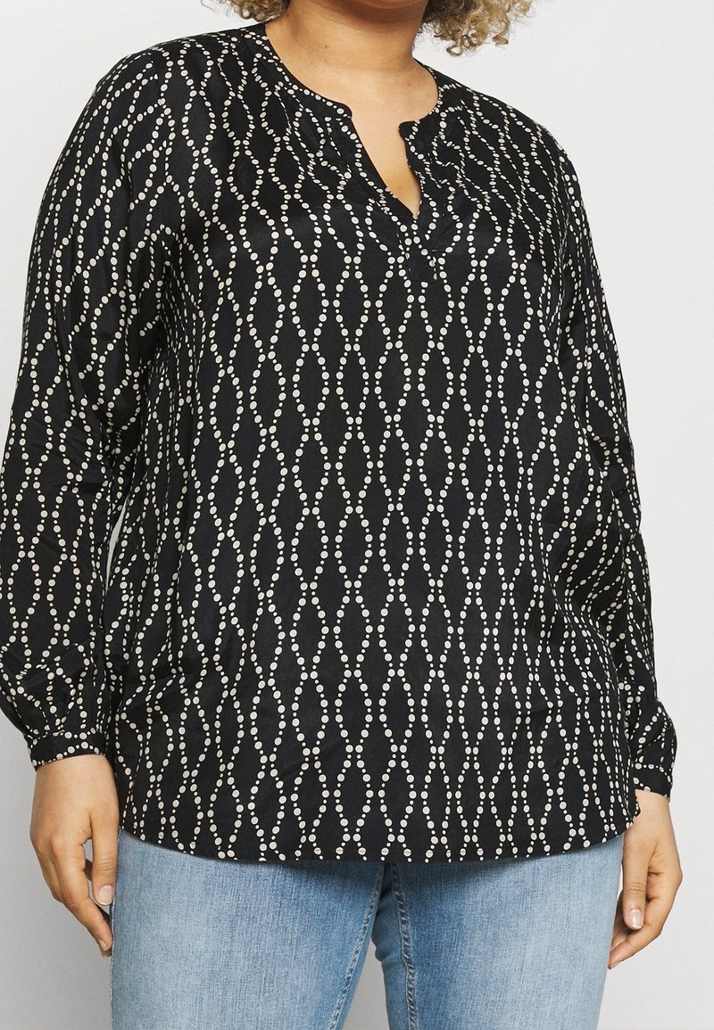 Black long-sleeve blouse with white dotted diamond pattern worn with light blue jeans by a person with curly hair.