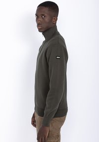 Mørkegrøn zip-up sweater med ribbede manchetter og kant. Fremstillet af struktureret strikmateriale, har den et lille logo-patch på venstre skulder.