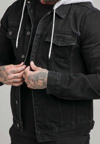 Man met getatoeëerde handen die een zwarte spijkerjack met grijze capuchon en witte trekkoorden dichtknoopt, met een zwart shirt eronder.
