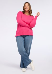 Donna sorridente che indossa un maglione rosa brillante, jeans a gamba larga blu e scarpe da ginnastica bianche, in piedi con le gambe incrociate su uno sfondo bianco.
