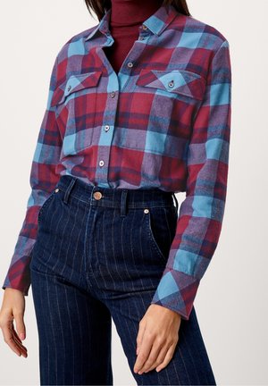 Vrouw draagt een roodblauw geruit overhemd over een bordeauxrode coltrui, gecombineerd met een high-waisted donkerblauwe jeans met krijtstreep.