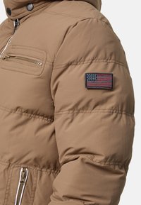 Beige Puffermantel mit vorderen Reißverschlusstaschen und einem aufgenähten amerikanischen Flaggenpatch auf dem oberen linken Ärmel.