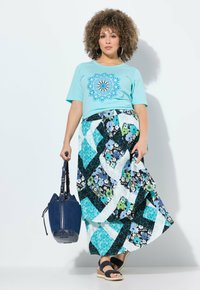 T-shirt turchese con stampa mandala, abbinata a una gonna maxi A-line multicolore in blu, nero e fantasie floreali. Porta con sé una borsa blu.