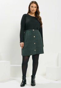 Schwarzes Langarmoberteil kombiniert mit einem minirock im grünen Karomuster, der mit einer Knopfleiste vorne geschlossen wird. Das Model trägt schwarze Strumpfhosen und Ankle Boots.