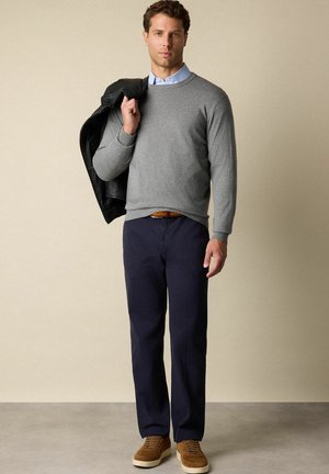 Muž v šedém svetru přes světle modrou košili, tmavě modrých kalhotách a hnědých botách, držící černou bundu přes rameno, stojící na jednobarevném pozadí.