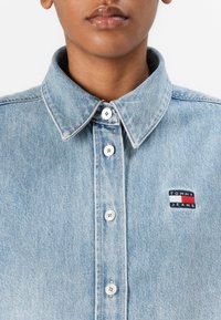 Ljusblå denimskjorta med spetsig krage, silverfärgade knappar och en liten broderad "TOMMY JEANS" logotyp på bröstet.