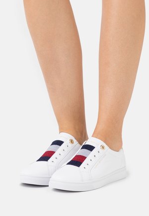 Zapatos blancos tipo slip-on con un diseño de rayas en tela azul marino y roja en la parte frontal, con detalles dorados cerca de los ojales y suelas texturizadas.