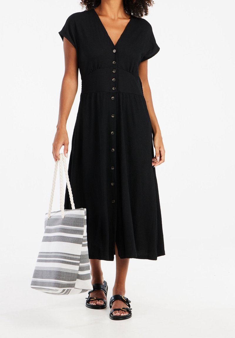 Femme portant une robe midi noire à boutons sur le devant, manches courtes, tenant un sac cabas rayé et portant des sandales noires à boucle.