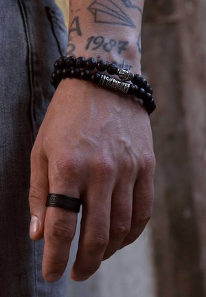 Black bead bracelet with a silver accent and a textured black ring on a hand with visible veins. Background features are blurred.