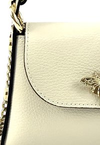 Beige lederen handtas met een gestructureerde afwerking, afgeronde flap en gouden vlinderaccent. Voorzien van een kettingriem en contrasterende zwarte randen.