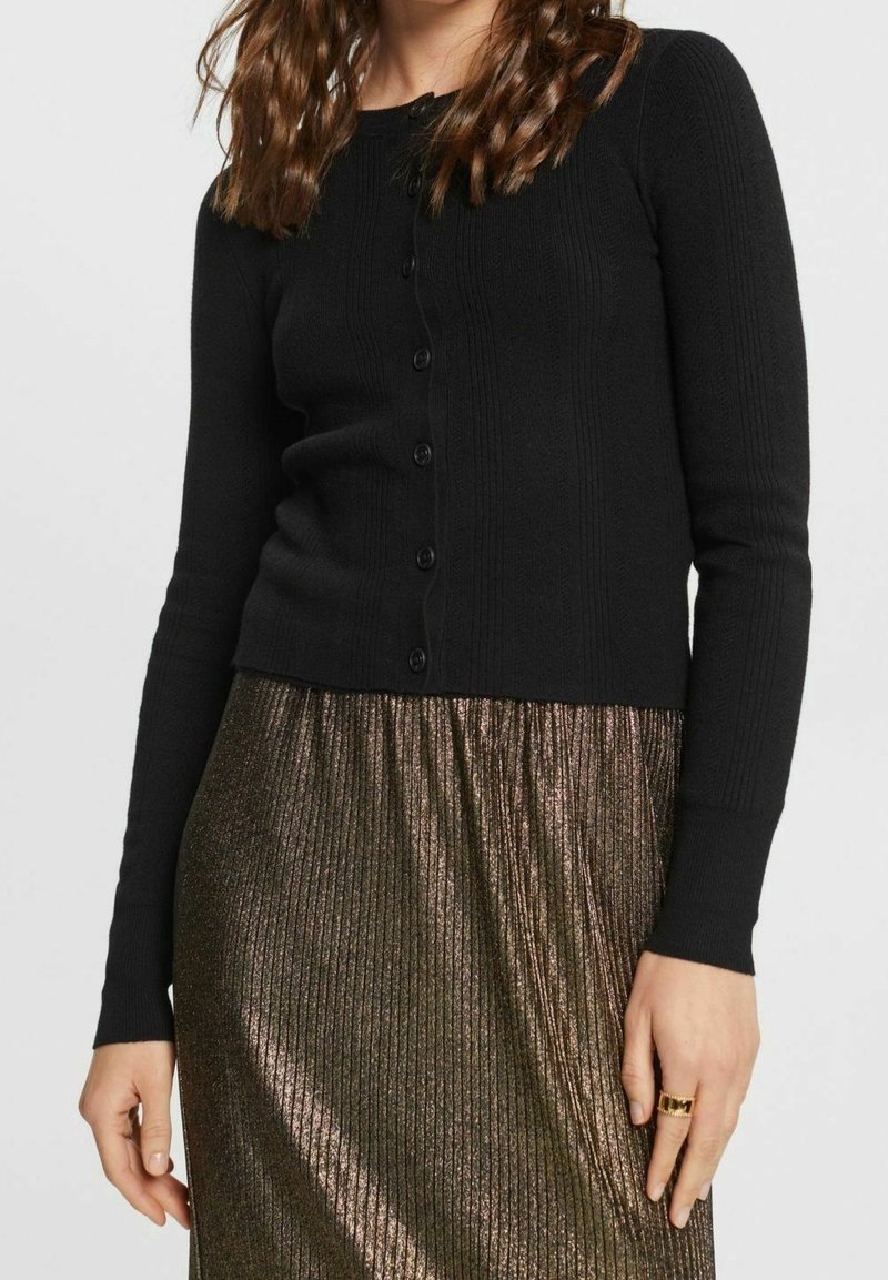 Zwarte geribbelde cardigan met lange mouwen en ronde hals, voorzien van een knoopsluiting aan de voorkant en een cropped zoom, gecombineerd met een metallic geplooide rok.