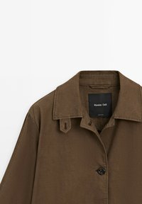 Hnědá bunda se strukturovaným límcem, zapínání na jeden knoflík a dvěmi významnými ramenními klapkami. Vyrobena z texturované tkaniny, minimalistický design.