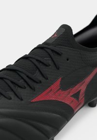Close-up de uma chuteira de futebol preta com parte superior texturizada e logótipo vermelho da Mizuno na lateral, apresentando cadarços pretos e sola com cravos.