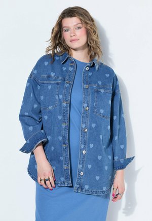 Vrouw met golvend haar die een blauwe spijkerblouse met hartjespatroon draagt over een blauwe jurk, staand tegen een effen lichte achtergrond.