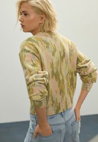Pulover tricotat în model de camuflaj verde și roz; are manșete și tiv ondulate, cu o croială lejeră și mâneci rulate, asortat cu blugi din denim.