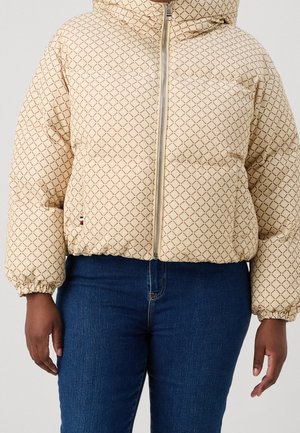 Osoba ubrana w beżową pikowaną kurtkę z brązowym wzorem w kratę i zamkiem błyskawicznym, w połączeniu z ciemnoniebieskimi jeansami.