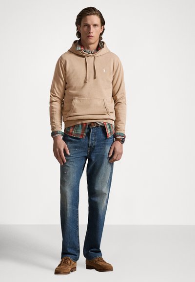Sweatshirt bege com bolso frontal, usado por cima de uma camisa xadrez. Combinado com jeans azuis desbotados e sapatos de camurça castanhos. Design simples e ajuste confortável.