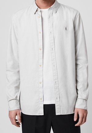 Mann trägt ein hellgraues, zum Knöpfen geeignetes Hemd über einem weißen T-Shirt und schwarzen Hosen, mit einem kleinen, gestickten Logo auf der Brust.