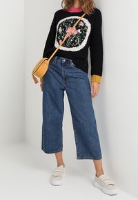 Kobieta ubrana w czarny sweter w kwiatowy wzór, szerokie niebieskie dżinsy sięgające do kostek, białe sneakersy i małą torebkę na ramię w kolorze musztardowym.