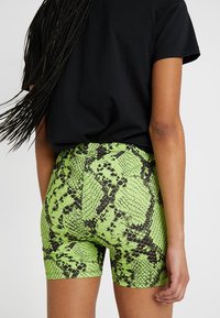 Persona con cabello largo trenzado, vistiendo una camiseta negra y pantalones cortos verdes con estampado de serpiente, vista desde atrás sobre un fondo liso.