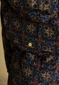 Melnā audumā ar ģeometrisku rakstu, kas ietver zeltainus rīkus, zilo akcentu un sarkano līniju, pabeigts ar apaļu zelta pogu detaļu.