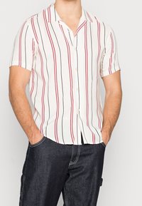 Camicia a maniche corte bianca con righe verticali rosse e nere. Presenta un colletto, abbottonatura frontale e una vestibilità rilassata. Indossata con pantaloni di denim scuri.
