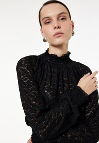 Sort blonde bluse med høj rynket hals og lange ærmer. Har teksturerede mønstre og samling detaljer på ærmeafslutningerne. Glat finish.