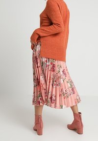 Maglione arancione con texture e orlo a coste, abbinato a una gonna a pieghe rosa con motivi floreali. Stivali alla caviglia rosa completano l'outfit.