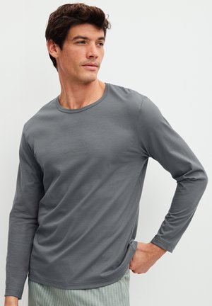 Mann trägt ein schlichtes graues Langarmshirt und hell gestreifte Hose, schaut zur Seite und hat eine Hand in der Hüfte.