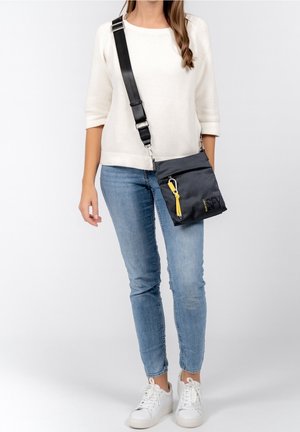 Nainen, joka pukeutunut valkoiseen villapaitaan, sinisiin farkkuihin ja valkoisiin tennareihin, kantaa mustaa olkalaukkua, jossa on säädettävässä hihnassa keltainen avaimenperä.