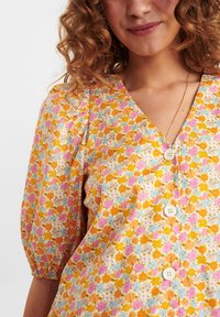 Blusa floral de botão em amarelo, com flores em rosa e azul, mangas curtas bufantes e decote em V. O tecido tem uma textura suave.