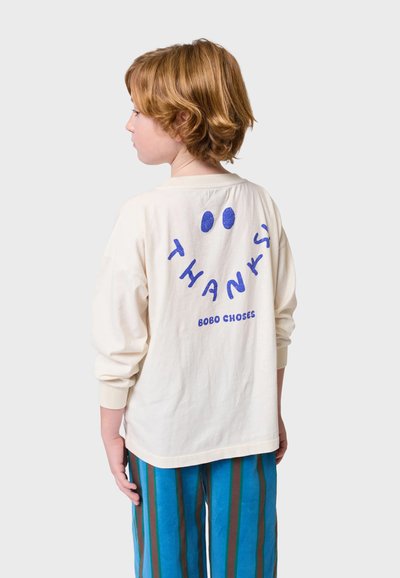 Košulja dugih rukava krem boje s plavim natpisom "HVALA" i dvije točke na leđima. Uparen s plavim prugastim hlačama.