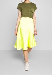 Olivově zelené tričko s ohrnutými rukávy a kapsou, doplněné jasně žlutou sukní s asymetrickým lemem. Na nohou bílé tenisky.