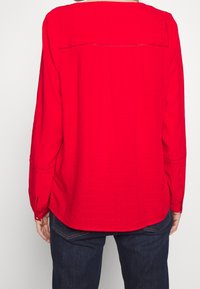 Blouse rouge à manches longues avec un tissu texturé, un ourlet arrondi et des broderies décoratives subtiles au niveau du plastron et des poignets.