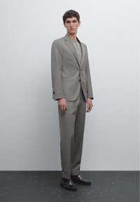 Grauer Anzug mit strukturierter Silhouette, Zweiknopfjacke, zulaufenden Hosen, strukturiertem hellgrauem Unterhemd und schwarzen Lederschuhen.