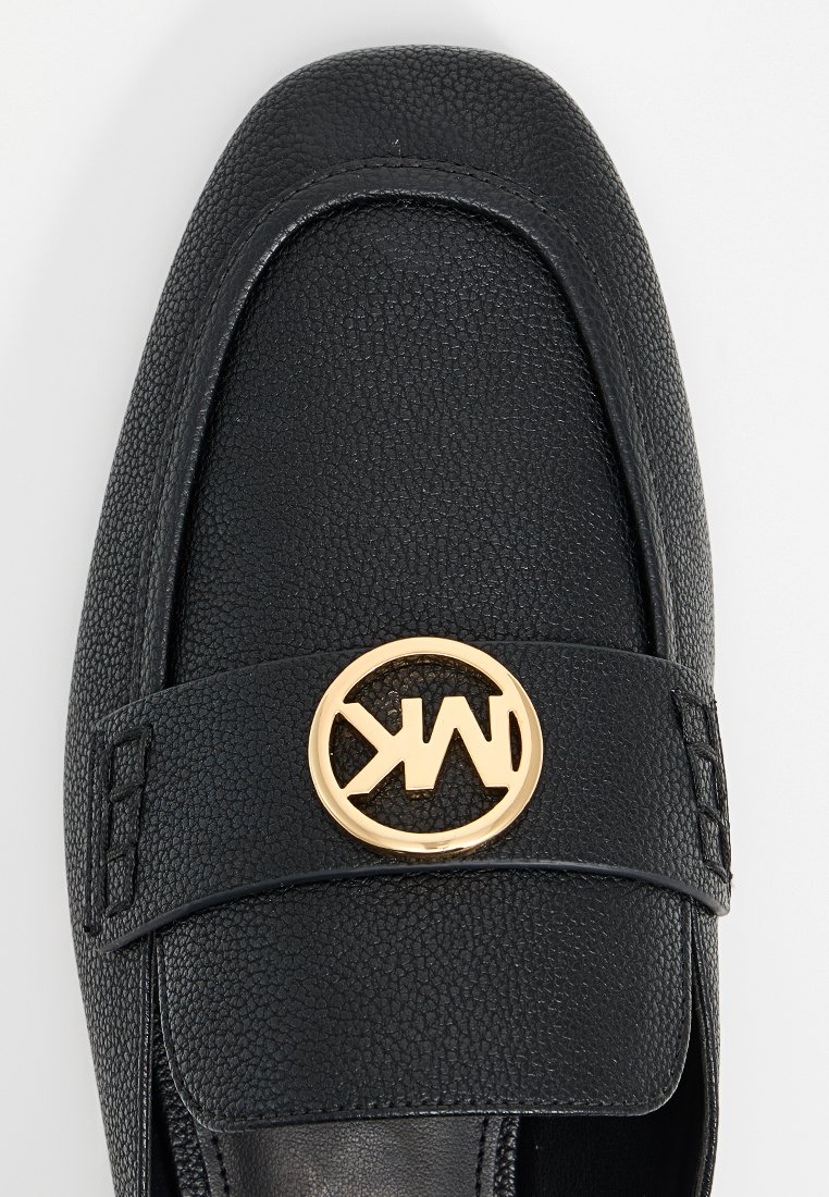 Zwarte leren instapper met een gestructureerde afwerking, voorzien van een gouden cirkelvormig logo-accent op de band. Vierkante neusontwerp met minimale stikseldetails.