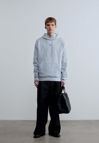 Ανοιχτό γκρι φούτερ με κουκούλα, μπροστινή τσέπη και κορδόνια, συνδυασμένο με μαύρο παντελόνι φαρδιάς γραμμής και μαύρη τσάντα. Ομαλή υφή υφάσματος.