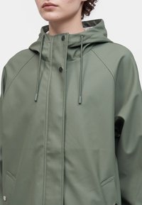 Personne portant un imperméable vert à capuche avec cordons et boutons-pression, montrée de la poitrine au menton sur un fond uni.