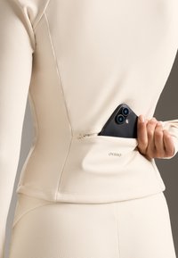 Long-sleeve beige top with textured fabric and a zippered pocket, holding a smartphone. Brand name "Oysho" is printed near the hem.