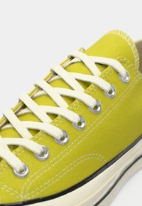 Adidași din pânză de un galben viu, cu sireturi albe, ochi de metal și talpă din cauciuc negru contrastant. Material texturat cu cusături vizibile.