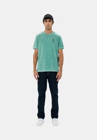 T-shirt verde menta a maniche corte, colletto rotondo e un piccolo logo nero; abbinato a jeans scuri e sneakers bianche.