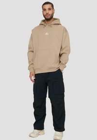 Felpa beige con tasca frontale e cappuccio con coulisse, abbinata a pantaloni cargo blu navy con multiple tasche. Scarpe da ginnastica completano il look.