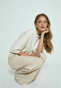 Kremowy, oversize'owy sweter z wysokim kołnierzem, w połączeniu z luźnymi, beżowymi spodniami. Modelka ma długie, falujące włosy i asymetryczne kolczyki.